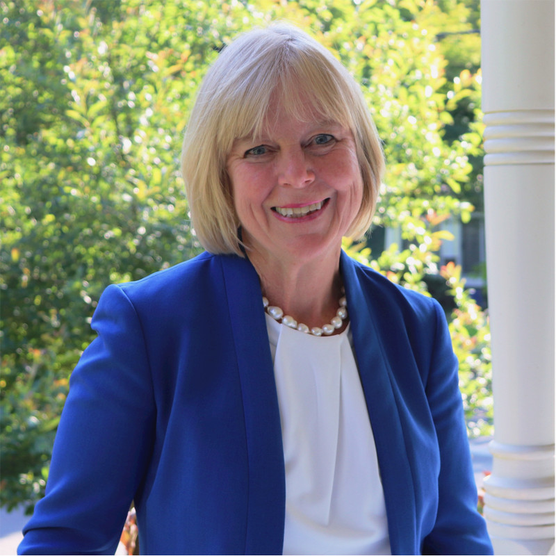 woman with short white hair, smiling and wearing a navy blue jacket with white shirt. she's outside, sitting on a porch on a bright sunny day